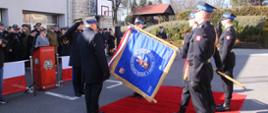 Zdjęcie przedstawia funkcjonariuszy w galowych mundurach salutujących przy sztandarze podczas ceremonii na czerwonym dywanie przed budynkiem komendy. W tle widoczny jest budynek jednostki z jasną elewacją i częścią z kamiennej cegły, zgromadzeni goście, drzewa oraz bezchmurne niebo.