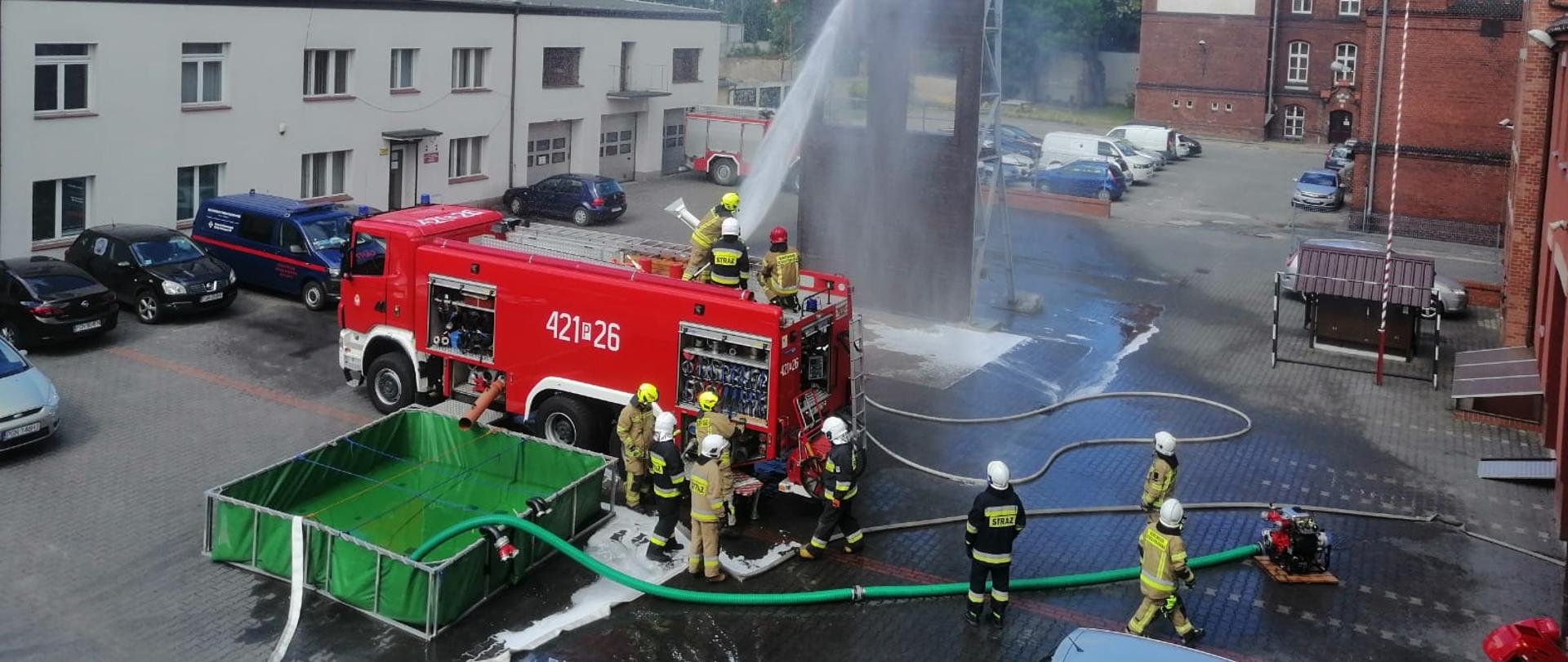 Na zdjęciu widać czerwony samochód strażacki z którego podawana jest woda przy użyciu działka wodnego znajdującego się na dachu pojazdu. Działko obsługuje trzech strażaków. Obok samochodu stoi zielony zbiornik wodny. Obok samochodu znajdują się ćwiczący strażacy