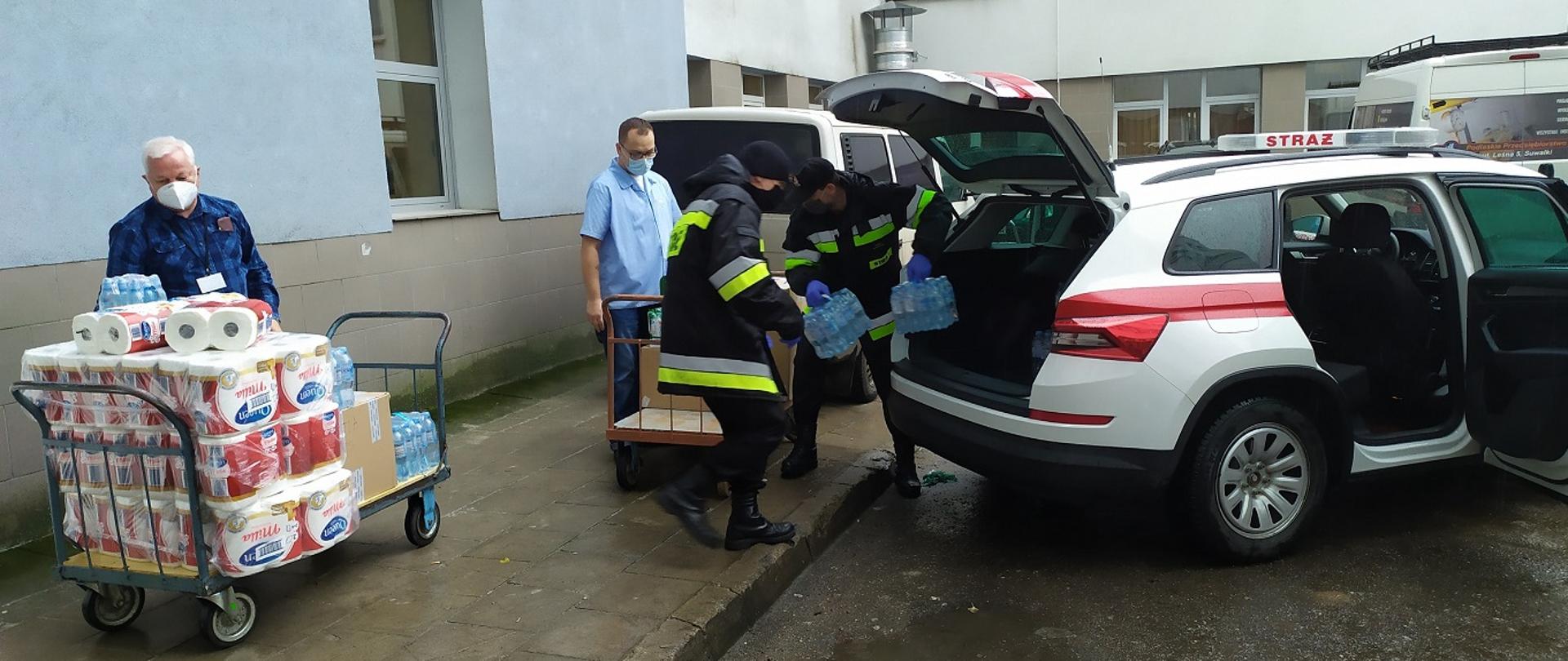 Na pierwszym planie paleta z ręcznikami papierowymi i pracownik szpitala. W głębi zdjęcia dwóch strażaków wyjmujących butelki z wodą z samochodu strażackiego, pracownik szpitala z wózkiem do przewozu przedmiotów oraz samochód dostawczy.
