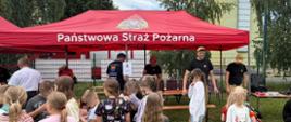 Na zdjęciu widać plenerowe wydarzenie edukacyjne zorganizowane przez Państwową Straż Pożarną. Pod czerwonym namiotem z napisem „Państwowa Straż Pożarna” strażacy rozmawiają z dziećmi i prezentują sprzęt ratowniczy. Przed namiotem zgromadzona jest grupa dzieci w kolorowych ubraniach, niektóre trzymają elementy wyposażenia strażackiego. W tle widoczne są drzewa, ogrodzenie oraz inne stanowiska służb. Scena przedstawia rodzinny, edukacyjny charakter wydarzenia, promującego bezpieczeństwo i pracę strażaków.