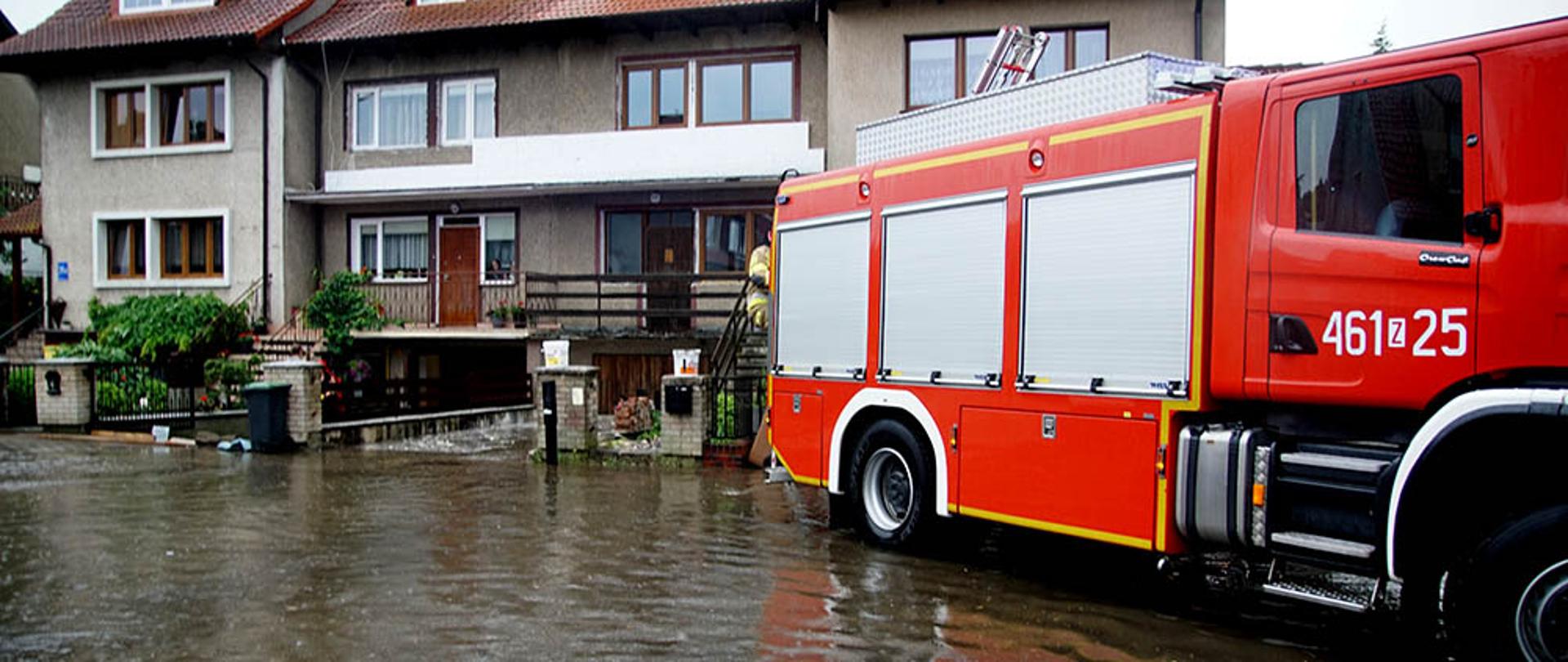Zdjęcie przedstawia wóz straży pożarnej na zalanej wodą ulicy.w tle widać zabudowania.