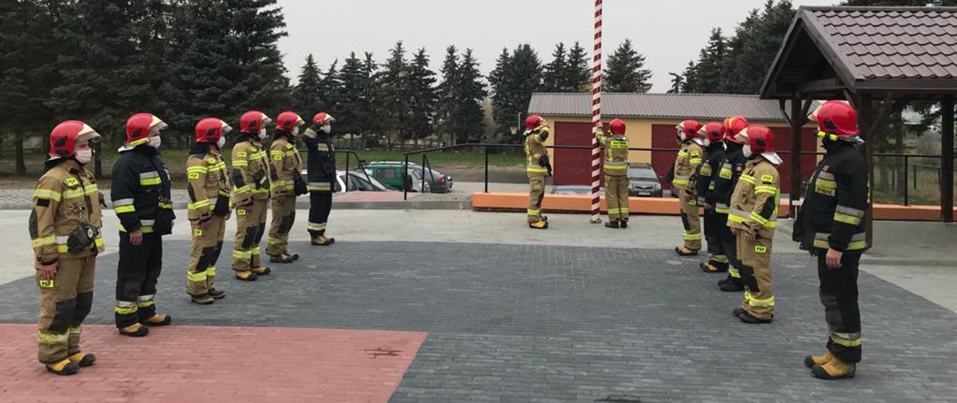 Funkcjonariusze w umundurowaniu koloru czarnego oraz piaskowego, w hełmach czerwonych stoją na placu szaro czerwonym przed masztem flagowym wieszają flagę narodową, w tle garaże i pojazdy samochodowe 