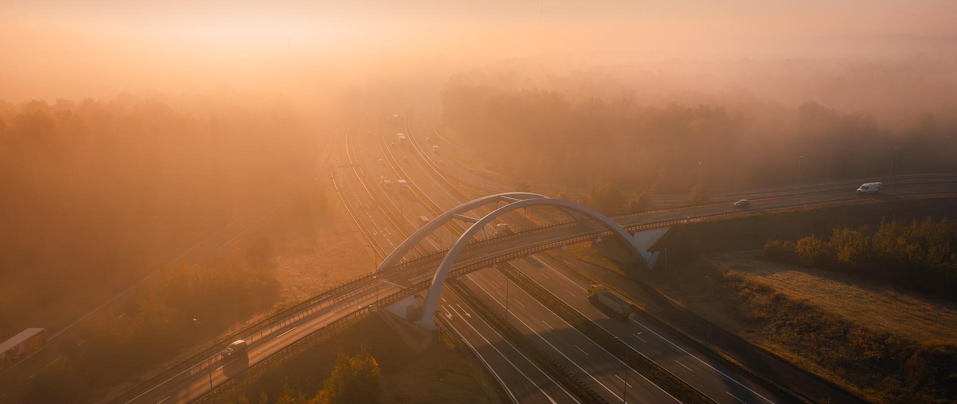 Widok z lotu ptaka na węzeł drogowy Murckowska w Katowicach spowity poranną mgłą. Na pierwszym planie widać autostradę A4 z kilkoma jadącymi pojazdami oraz charakterystyczny łukowy most nad jezdniami. W tle rozciąga się zalesiony teren, częściowo ukryty w gęstej, złocistej mgle, którą rozświetla wschodzące słońce.