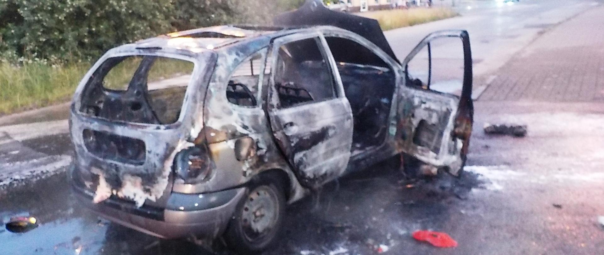 Na zdjęciu widzimy spalony samochód osobowy marki Renault Scenic. Dookoła pojazdu widać wodę, którą gaszony był pojazd. W tle widać jadący samochód po drodze w przeciwnym kierunku do stojącego spalonego auta oraz zabudowania.