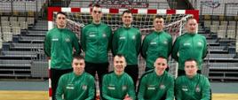 Zdjęcie przedstawia drużynę mężczyzn w zielonych dresach sportowych, ustawionych na hali sportowej przed bramką do piłki ręcznej. Dziewięciu zawodników pozuje do zdjęcia – pięciu stoi w tylnym rzędzie, a czterech klęczy z przodu. Wszyscy mają na sobie jednakowe zielone bluzy z herbem oraz logotypami sponsora lub organizacji. Zdjęcie zostało wykonane na tle pustych trybun w hali z widocznym logo klubu i obiektu sportowego. Atmosfera zdjęcia sugeruje profesjonalną lub półprofesjonalną drużynę sportową, prawdopodobnie związaną z piłką ręczną lub futsalem.