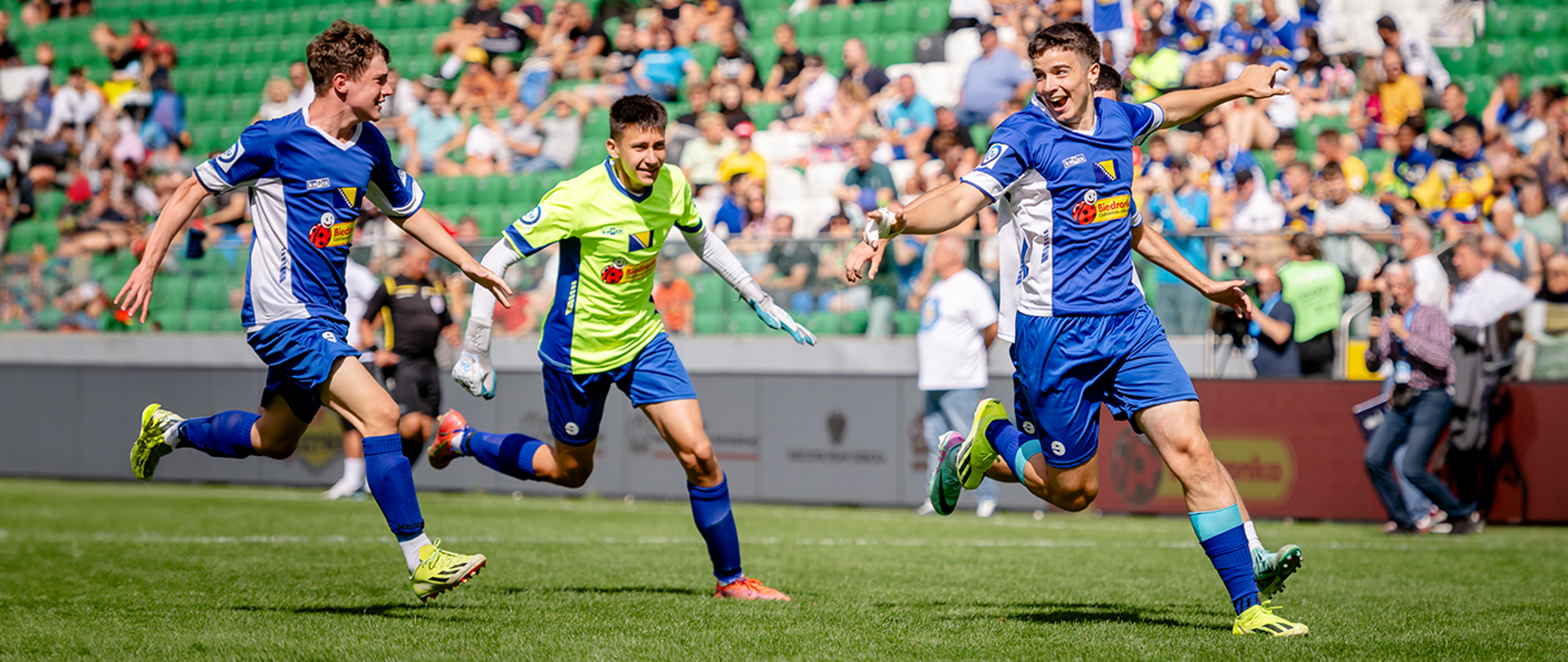 Trzech piłkarzy w niebieskich koszulkach biegnie razem, z szerokimi uśmiechami i rozłożonymi rękami, jakby świętowali ważny moment. W tle widać trybuny pełne kibiców. Radość na ich twarzach sugeruje, że być może właśnie odnieśli sukces w jednym z meczów turnieju.