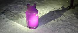 Różowa butla gazowa stojąca na śniegu nocą, oświetlona silnym światłem latarki; w tle ciemne podwórko i długi cień rzucany na śnieg. 