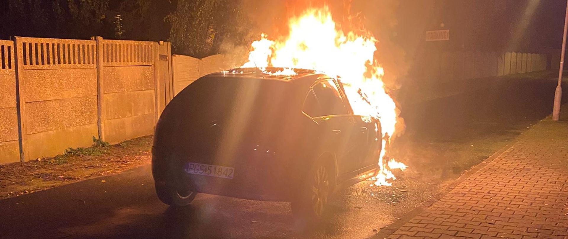 Zdjęcie przedstawia samochód osobowy w płomieniach, stojący na drodze.