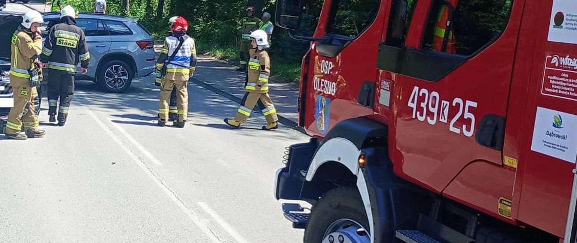 Na zdjęciu widać pojazd ratownniczy i strażaków podczas akcji, na drodze samochód po wypadku blokuje dwa pasy ruchu w oddali funkcjonariusze Policji.