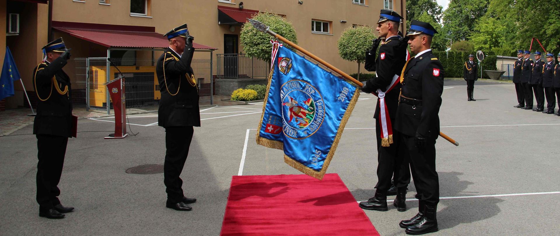 Na asfaltowym placu po prawej stronie stoi poczet sztandarowy z pochylonym sztandarem – naprzeciw niego dwóch strażaków w umundurowaniu galowym – salutują. Pomiędzy nimi na asfalcie czerwony dywan. Z tyłu po lewej stronie w szeregu stoi kilku strażaków w umundurowaniu galowym. W tle budynek Jednostki ratowniczo-gaśniczej.