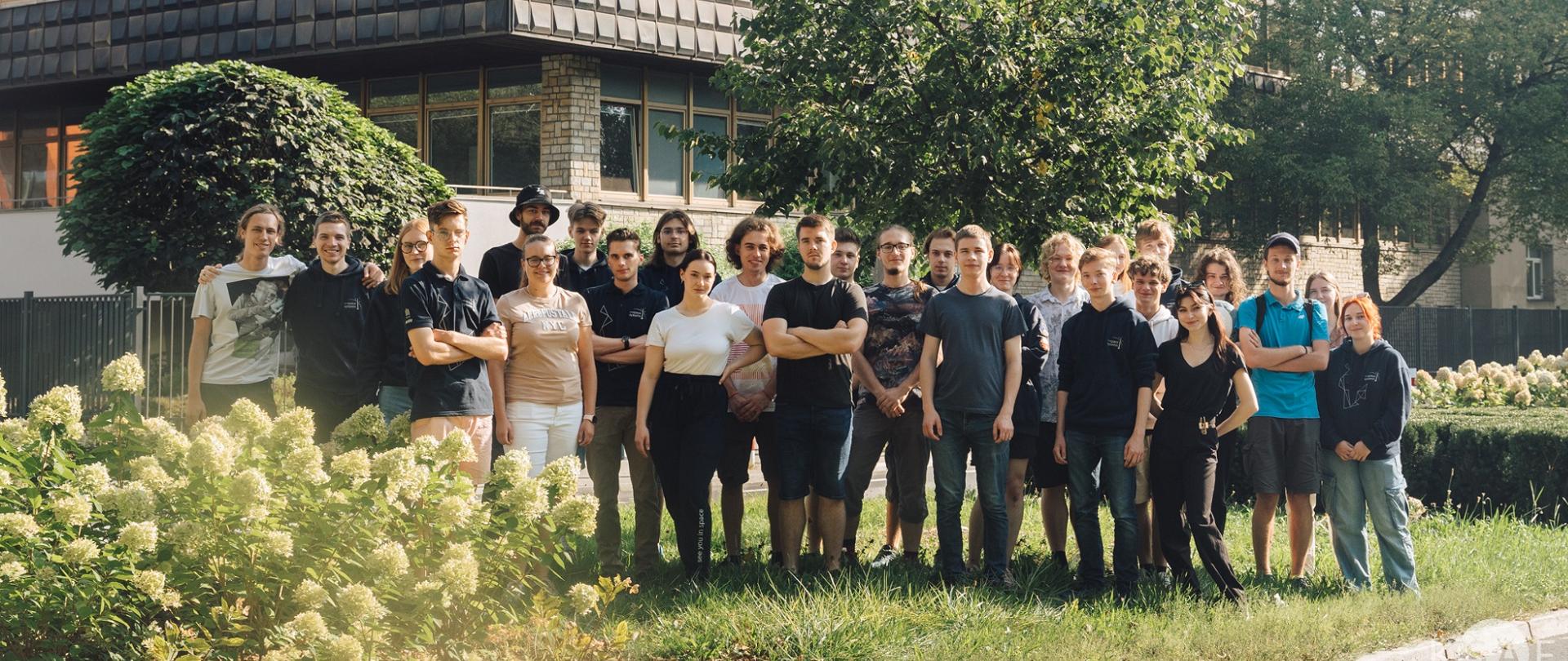 Grupa młodych ludzi stoi na trawie przed szarym kilkupiętrowym budynkiem.