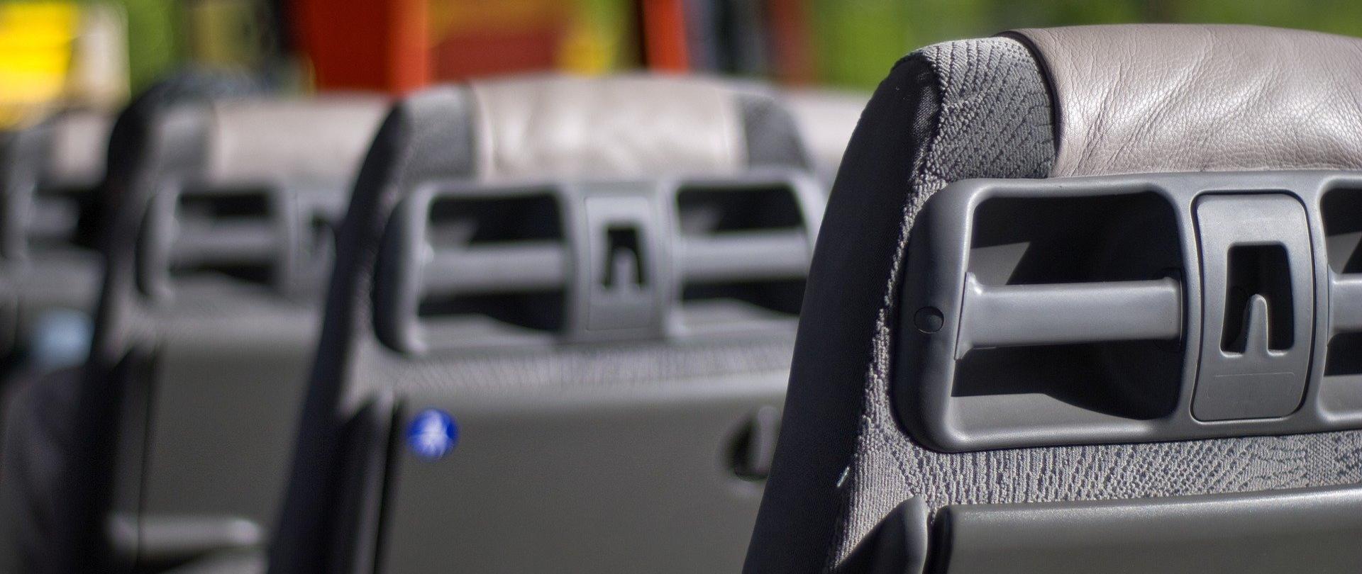 Zdjęcie przedstawia rząd foteli autobusowych. Ujęcie boczne. Na pierwszym planie - po prawej stronie jeden z nich. W tle widać okno autobusu i kolorową zasłonkę.