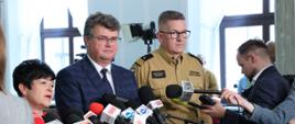 Osoby cywilne oraz funkcjonariusz Państwowej Straży Pożarnej stoją obok siebie przed mikrofonami.
