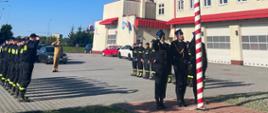 na placu przed Jednostka Ratowniczo Gaśnicza nr 2 stoją strażacy w dwóch szpalerach naprzeciw siebie na uroczystej zbiórce. Pomiędzy szpalerami stoi dowódca. Strażacy ubrani są w czarne ubrania koszarowe. Za szpalerami widoczny jest maszt na który poczet flagowy zawiesza biało-czerwona flagę