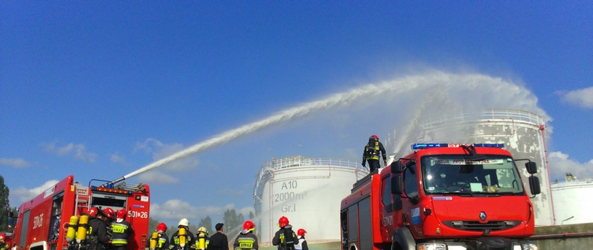 Zdjęcie przedstawia podawanie piany na zbiorniki z paliwem z działek wodno-pianowych samochodów