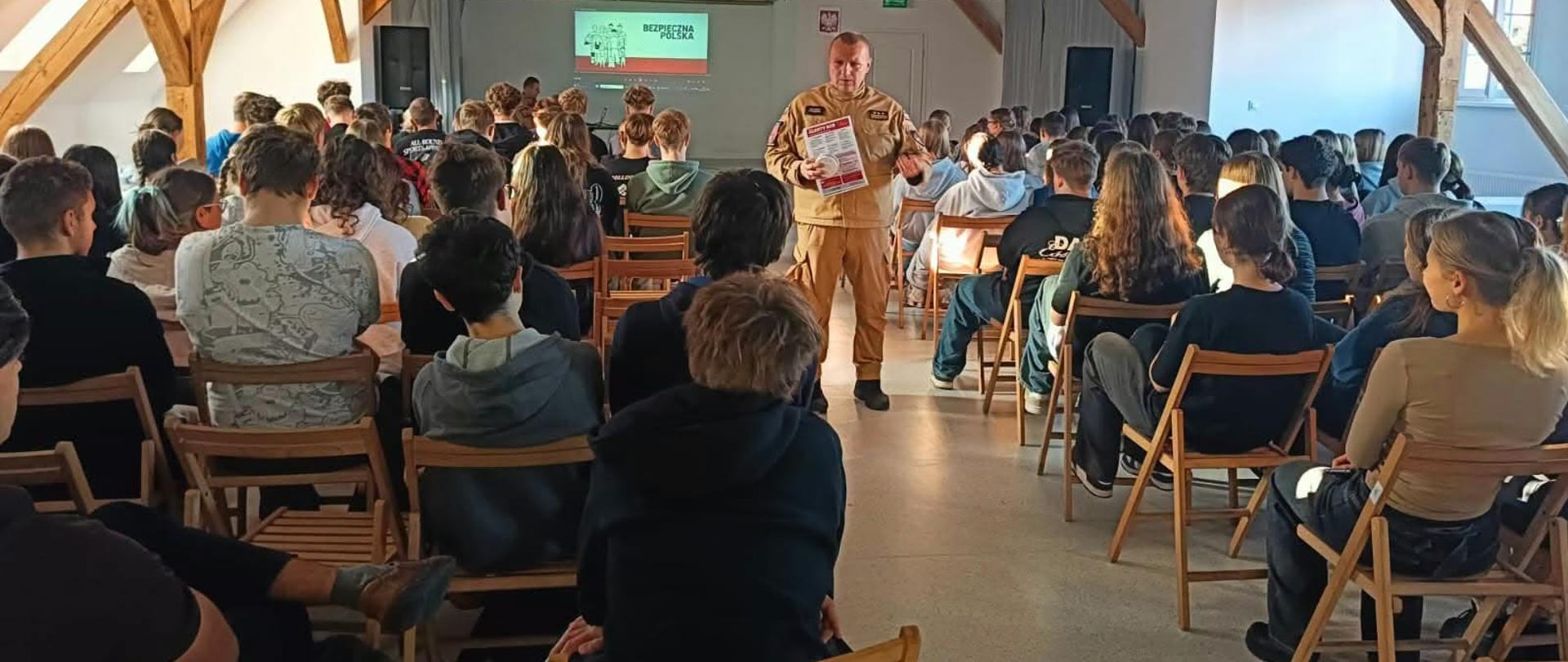 Na zdjęciu widać salę wypełnioną młodzieżą uczestniczącą w spotkaniu edukacyjnym. Pośrodku stoi strażak w mundurze, trzymający materiały informacyjne. W tle wyświetlana jest prezentacja „Bezpieczna Polska”.