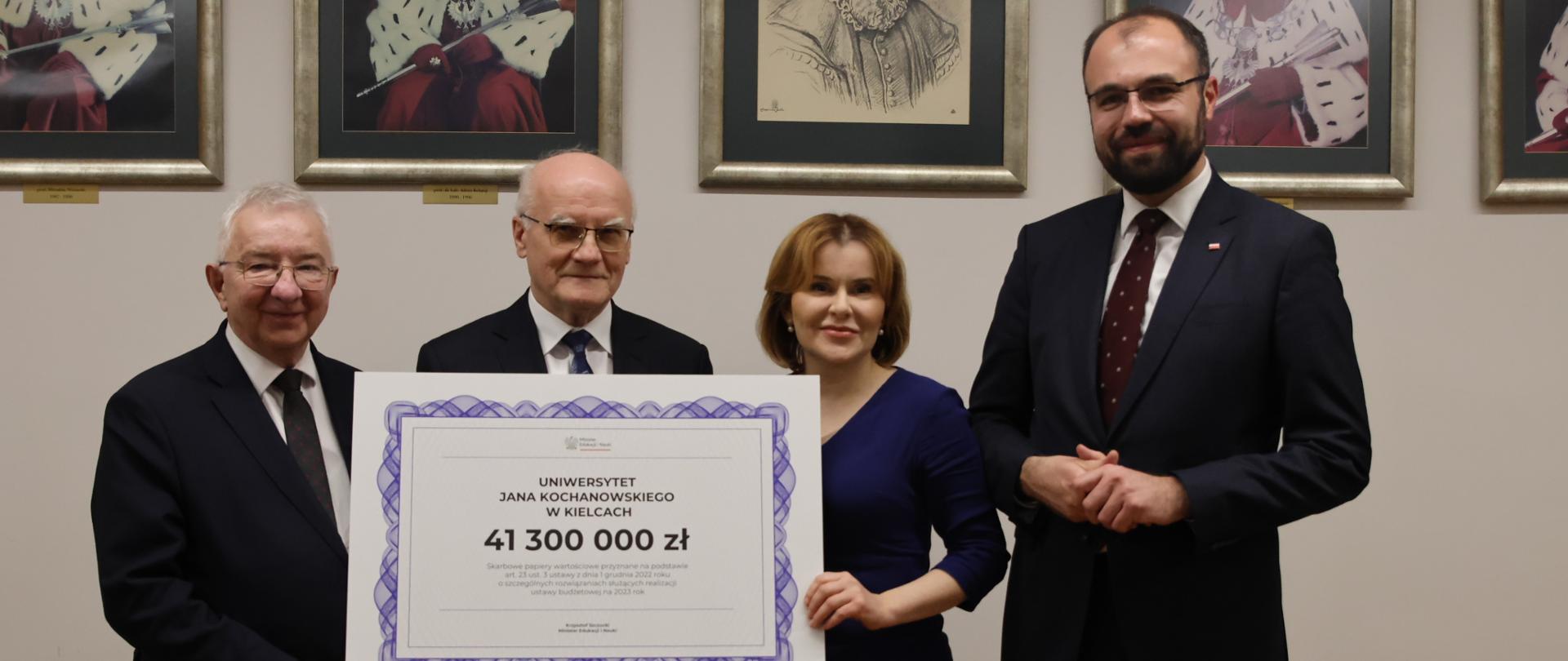 Dwaj mężczyźni w garniturach i kobieta trzymają wydrukowany czek z kwotą 41300000 zł. Obok stoi minister Szczucki. W tle na ścianie wiszą portrety rektorów. 