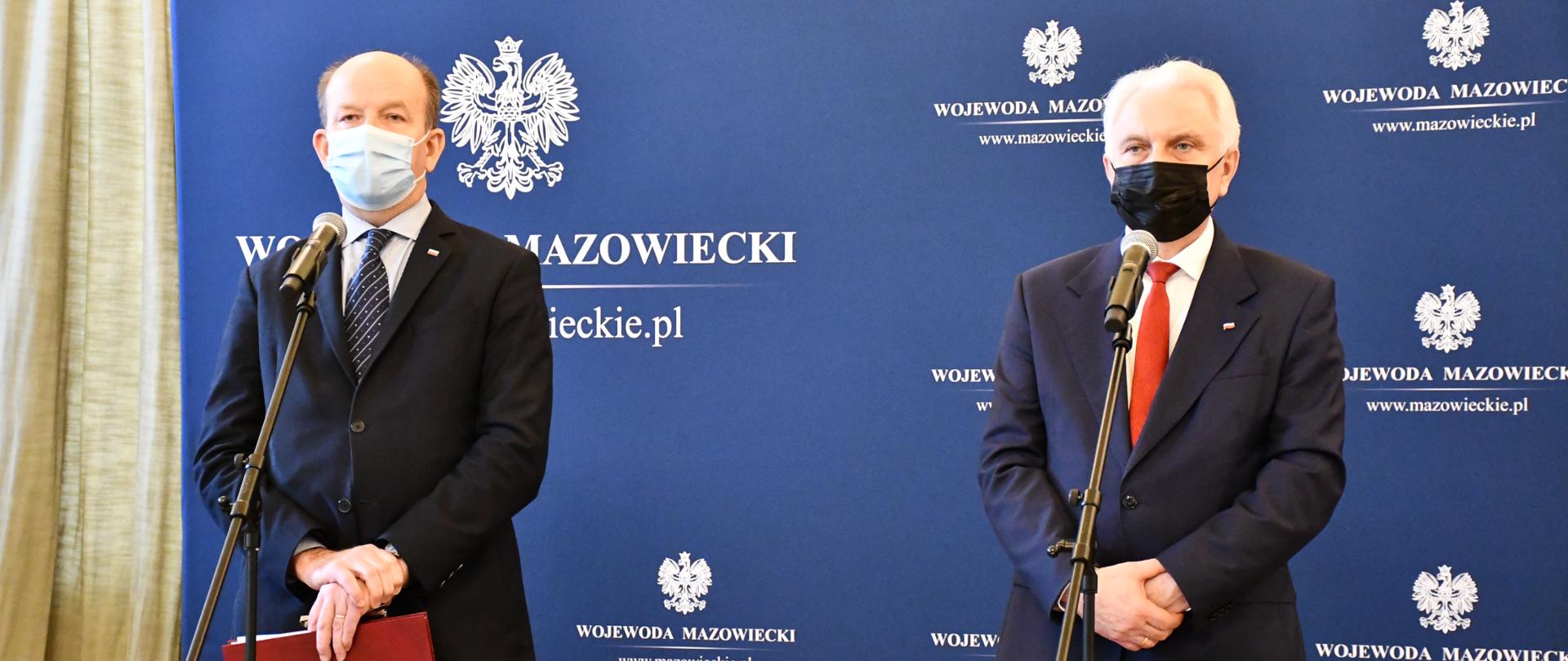 Na zdjęciu znajduje się dwóch mężczyzn, ubranych w garnitury. Stoją oni na tle granatowej ścianki wizerunkowej Wojewody, na której znajdują się różnej wielkości napisy "Wojewoda Mazowiecki" oraz Godło Polski. Mężczyźni Ci są w trakcie wystąpienia publicznego - stoją przed mikrofonami. 