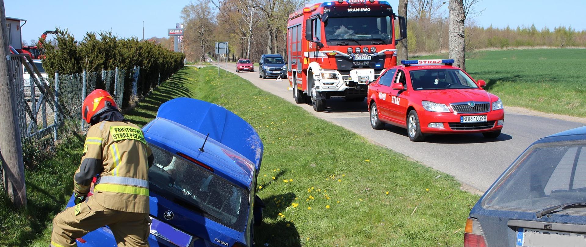 Z lewej strony zdjęcia na zielonym poboczu drogi stoi niebieskie auto przy nim strażak. Z prawej na drodze ustawione dwa samochody strażackie, czerwone.