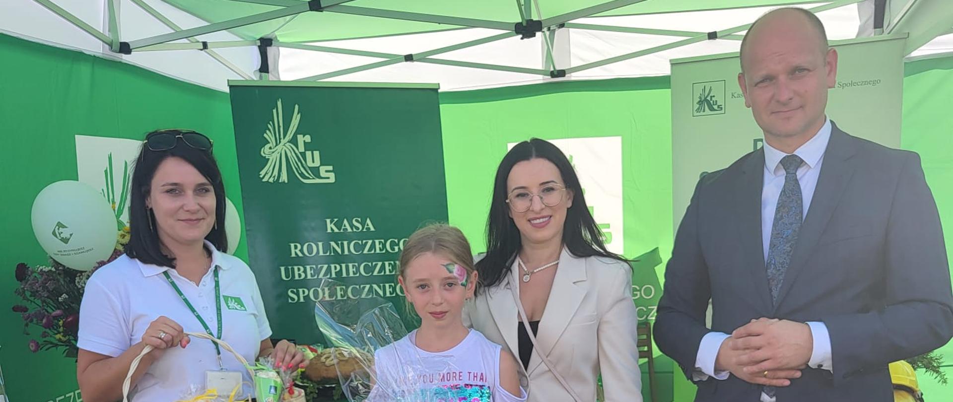 Na tle stoiska informacyjnego dwie kobiety, jeden mężczyzna oraz dziecko, które trzyma nagrodę zdobytą w konkursie.