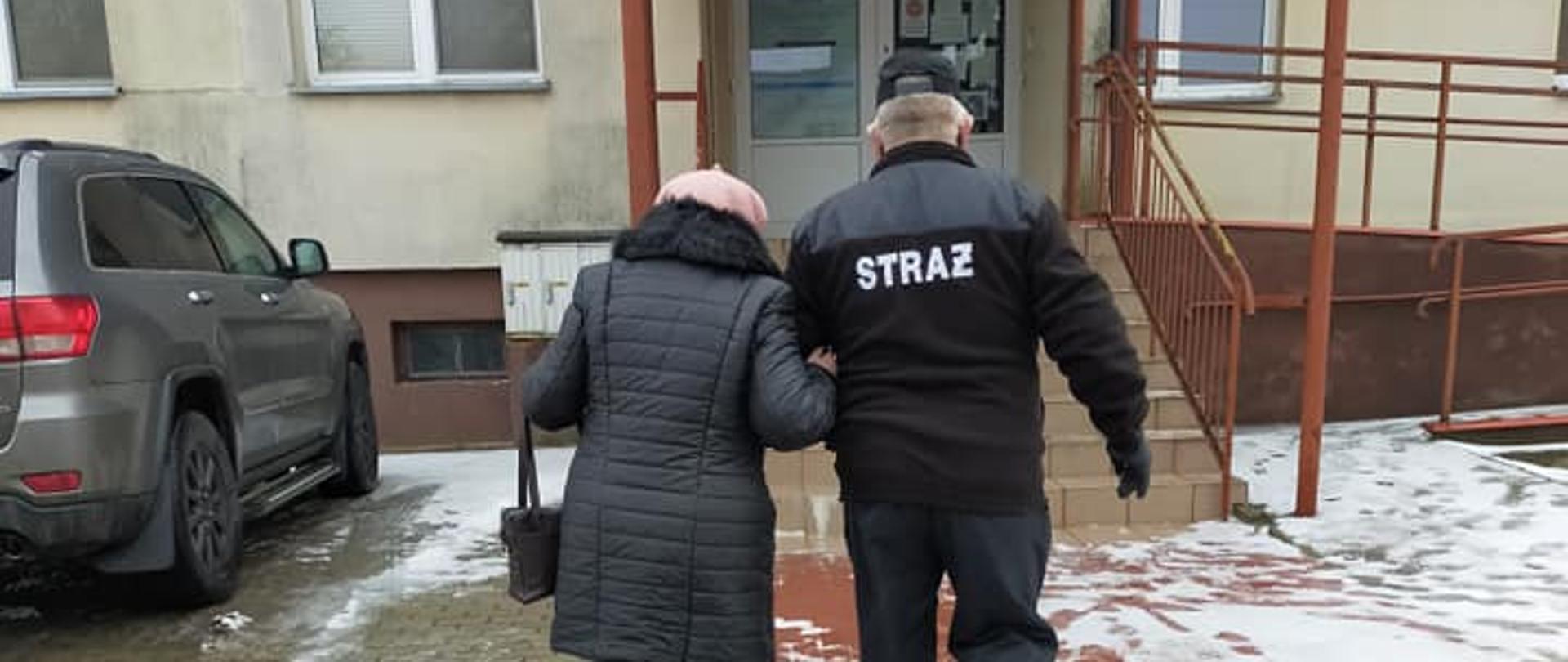 Na zdjęciu widać jak strażak OSP prowadzi starszą kobietę pod ramię do Ośrodka Zdrowia.