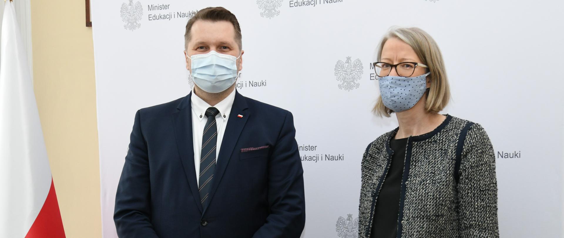 Minister Przemysław Czarnek wspólnie z Ambasador Zjednoczonego Królestwa Wielkiej Brytanii i Irlandii Północnej Anną Clunes. W tle flaga Polski