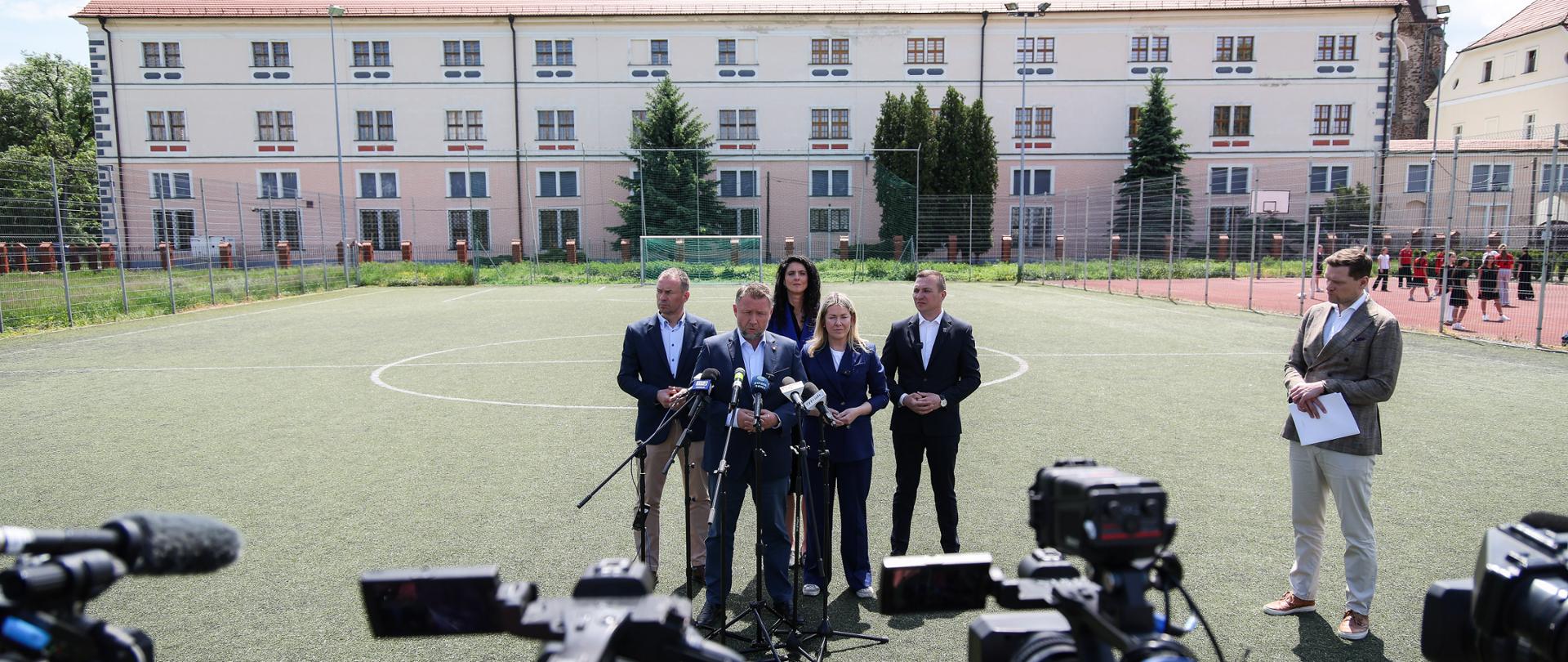 Konferencja prasowa. Uczestnicy stoją za mikrofonami na boisku. W tle budynek szkoły. Na pierwszym planie kamery.