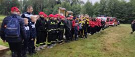 Grupa uczestników I Warsztatów Młodzieżowych Drużyn Pożarniczych „ISKRA XXVI” ustawiona w rzędzie na trawniku w Leśnictwie Goleszów. Młodzież w mundurach i dresach sportowych skupiona jest przed przystąpieniem do kolejnych zadań, a w tle widoczne są wozy strażackie.