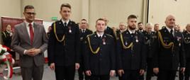 Z lewej Pan Wojewoda w siwym garniturze, obok niego wyróżnieni strażacy w granatowych mundurach wyjściowych bez nakryć głowy, z prawej Komendant Wojewódzki PSP. Wszyscy stoją w jednym rzędzie. Uroczystość przebiega w sali Urzędu. W tle pododdział strażaków. 