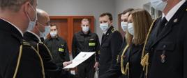 Komendant powiatowy mł. bryg. Arkadiusz Makowski ubrany w mundur galowy ze sznurem w maseczce ochronnej na twarzy wręcza awans strażakowi tutejszej komendy, w lewej dłoni trzyma teczkę na akta a prawą dłoń trzyma w uścisku strażaka, w głębi funkcjonariusze stoją w szeregu, na wprost trzech strażaków w pozycji zasadniczej ubranych w ubranie koszarowe w maseczkach ochronnych na twarzy