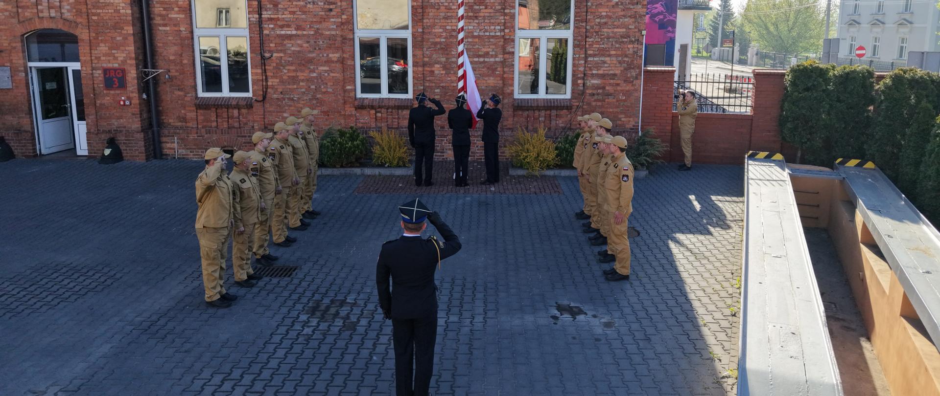 Na zdjęciu widać uroczystą ceremonię przy maszcie flagowym przed budynkiem z czerwonej cegły. Strażacy ustawieni są w dwóch równych szeregach naprzeciw siebie i oddają honor, salutując. W centrum, przy maszcie, trzech funkcjonariuszy w ciemnych, galowych mundurach podnosi flagę Polski. Na pierwszym planie stoi dowódca (również w mundurze galowym), który także salutuje, zwrócony w stronę flagi.