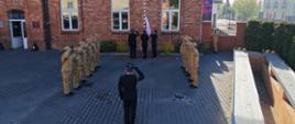 Na zdjęciu widać uroczystą ceremonię przy maszcie flagowym przed budynkiem z czerwonej cegły. Strażacy ustawieni są w dwóch równych szeregach naprzeciw siebie i oddają honor, salutując. W centrum, przy maszcie, trzech funkcjonariuszy w ciemnych, galowych mundurach podnosi flagę Polski. Na pierwszym planie stoi dowódca (również w mundurze galowym), który także salutuje, zwrócony w stronę flagi.