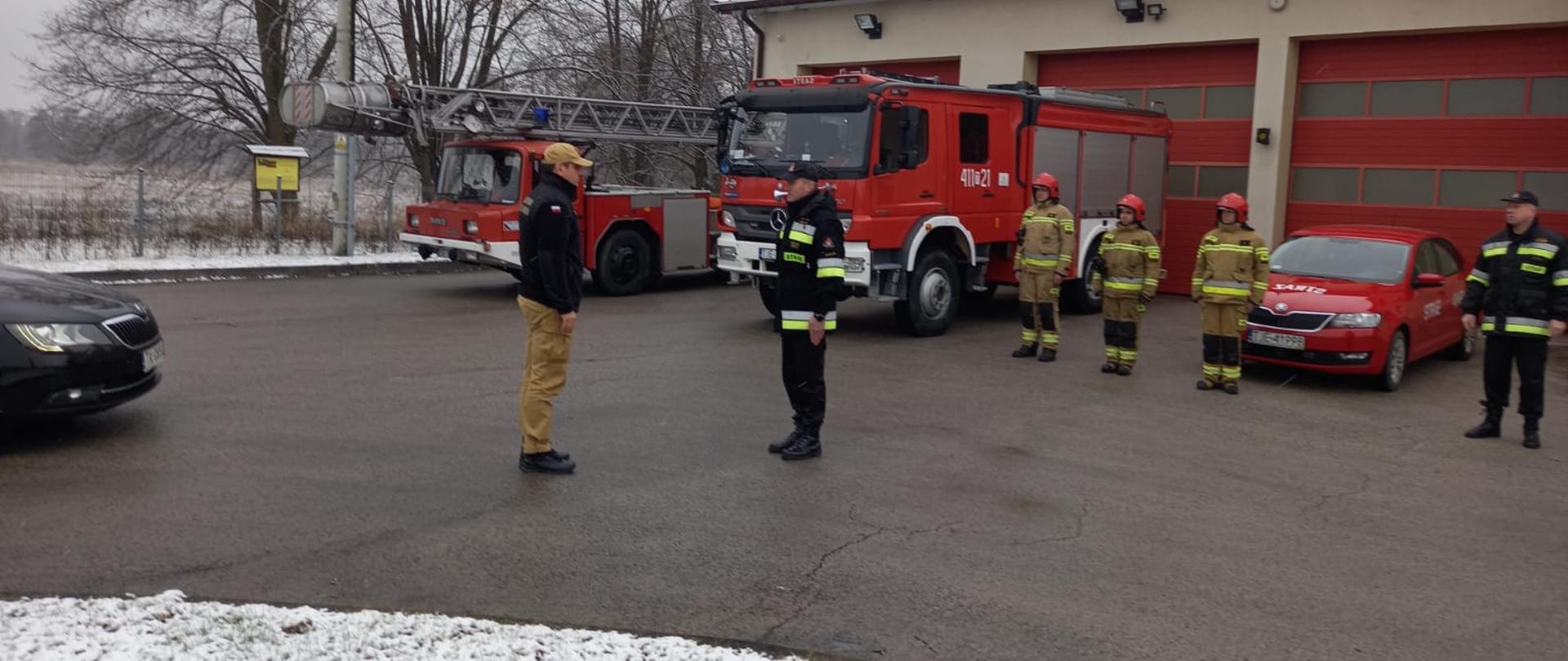 Zdjęcie przedstawia świętokrzyskiego komendanta wojewódzkiego PSP w Kielcach st. bryg. Krzysztofa Cioska, podczas składania meldunku przed dowódcę Jednostki Ratowniczo-Gaśniczej w Jędrzejowie. Za ich plecami ustawione dwa samochody ratowniczo-gaśnicze oraz strażacy w umundurowaniu specjalnym. Po prawej stronie widać komendanta powiatowego PSP w Jędrzejowie st. bryg. Pawła Jakubowskiego.