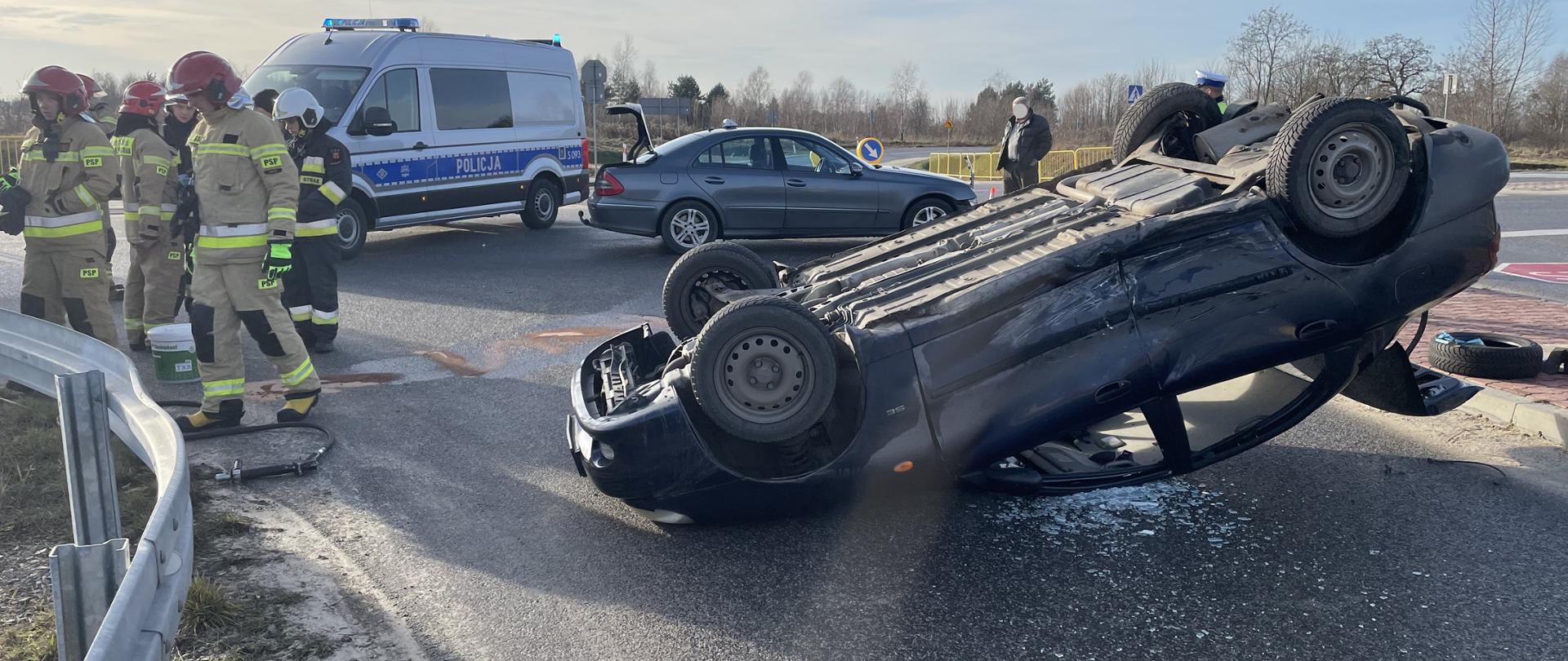 Na pierwszym planie widać samochód osobowy leżący na dachu na ulicy. Po lewej stronie grupa strażaków, a w tle od lewej radiowóz policyjny i drugi z uszkodzonych samochodów.