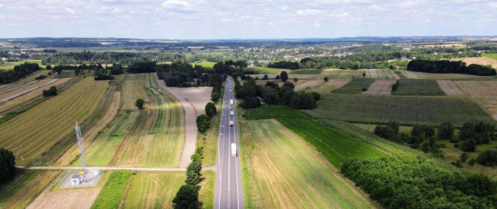 Droga biegnąca wśród pól, w oddali budynki, niebo zachmurzone