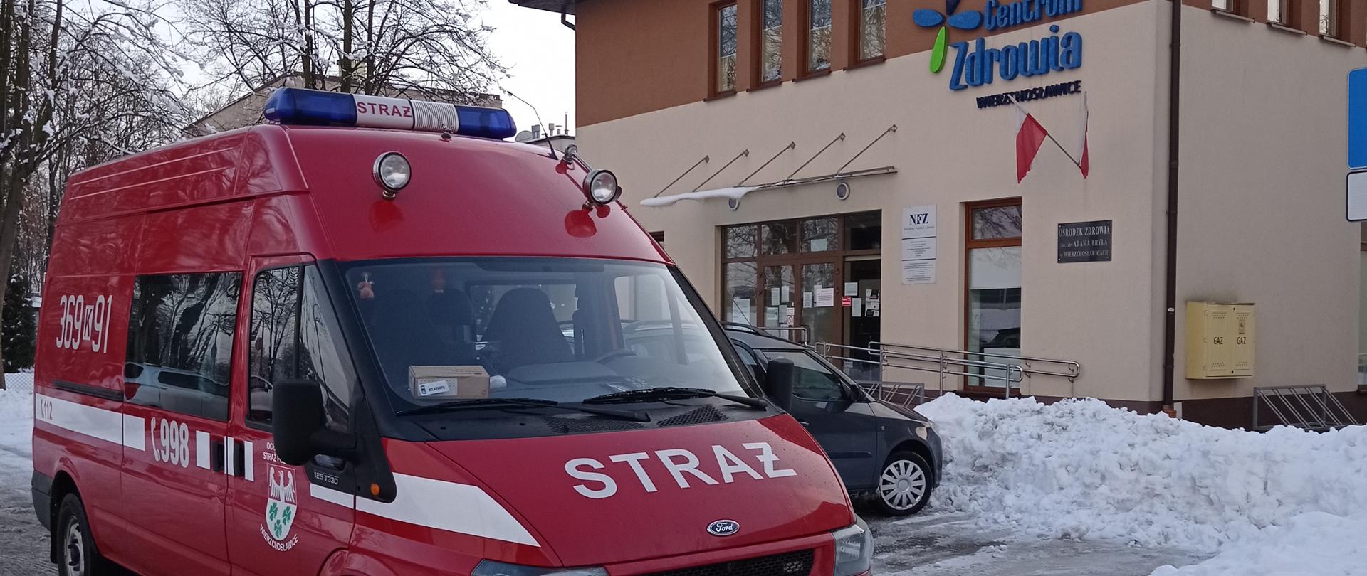 Na zdjęciu na ulicy stoi pojazd pożarniczy Ford Transit – przystosowany do przewozu osób. W tle (po prawej stronie) za pojazdem widać budynek - Ośrodek Zdrowia im. dr Adama Bryla w Wierzchosławicach. W otoczeniu widać śnieg usunięty z ulicy i chodnika.