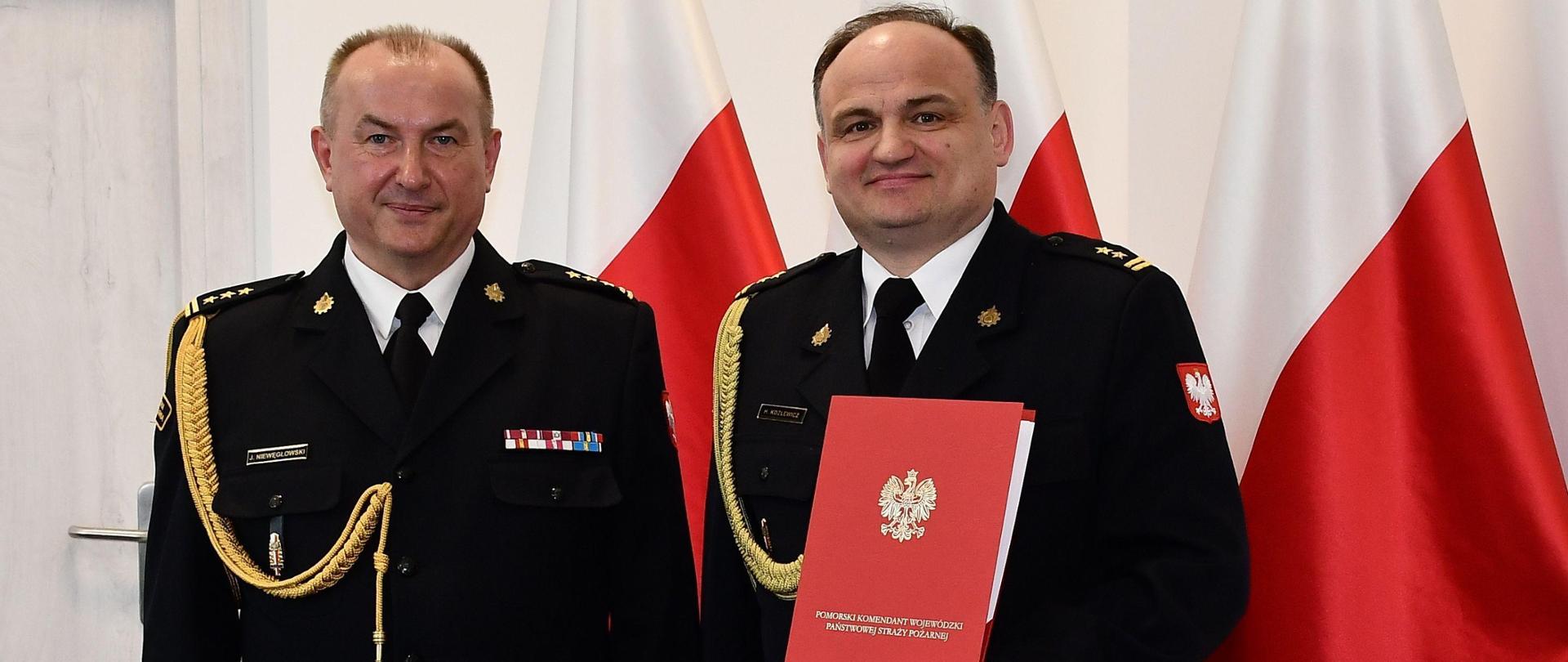 Dwóch strażaków w mundurach wyjściowych ze sznurem stoi obok siebie mężczyzna w środku jest bez teczki za nimi ustawione są trzy flagi Polski wisi obraz oraz znajdują się drzwi. 