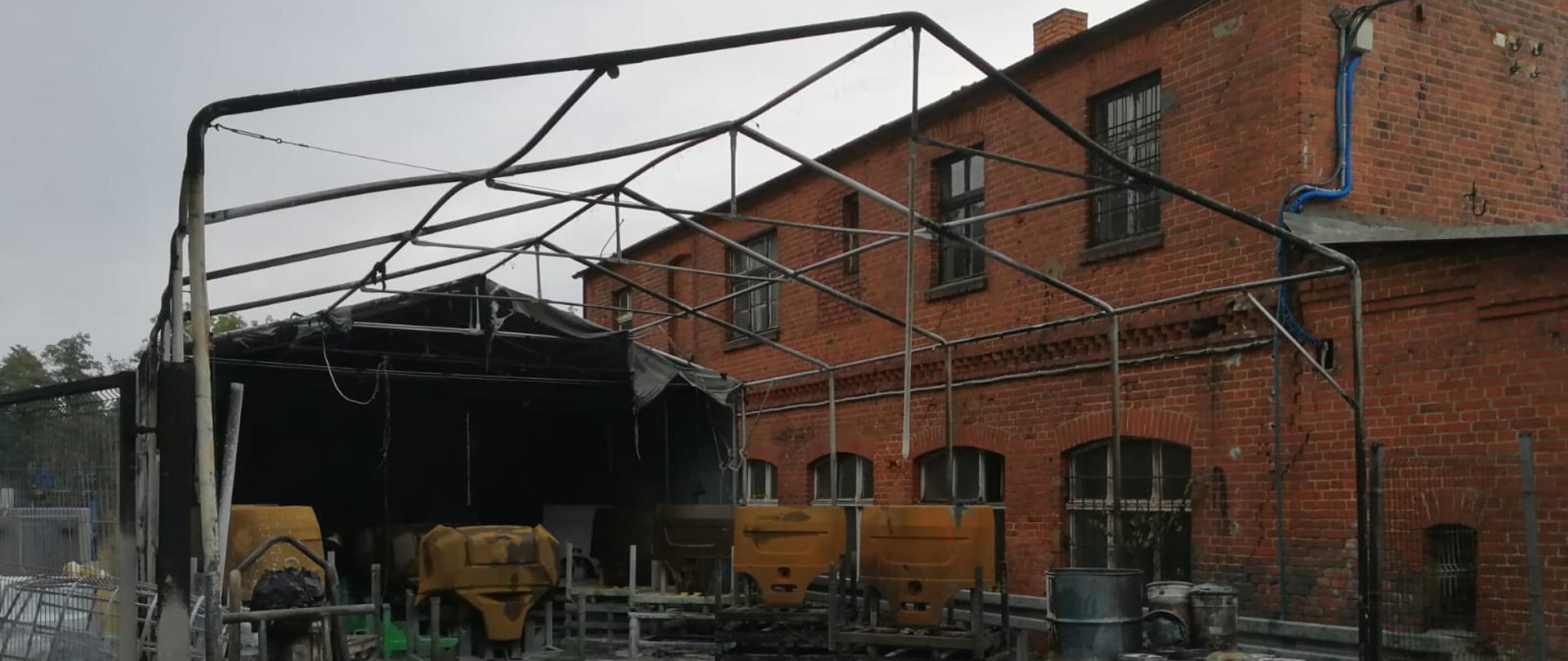 Na zdjęciu stelaż namiotu magazynowego. Pokrycie zostało całkowicie spalone. W tle budynek z czerwonej cegły.