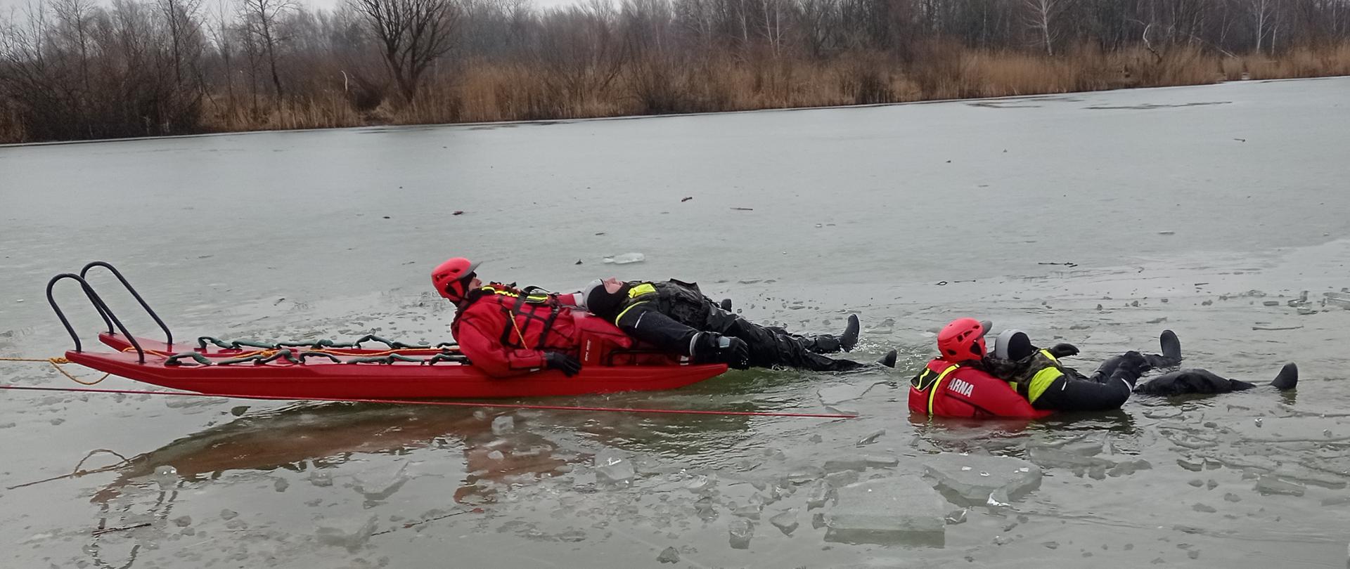 Na zdjęciu widać dwóch strażaków-ratowników na lodzie. Obaj ubrani są w czerwone skafandry i kaski. Jeden strażak leży na czerwonych plastikowych saniach i trzyma osobę wyciągniętą z wody, drugi jest w wodzie i trzyma osobę będącą w wodzie. Obie osoby ratowane przez strażaków ubrane są w czarne skafandry. W oddali widać zamarznięty akwen wodny i zalesiony brzeg.