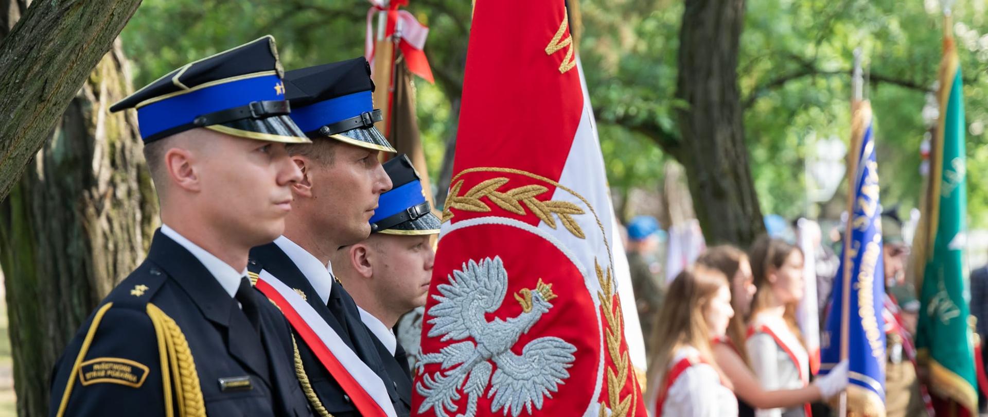 Z lewej poczet sztandarowy Komendy Miejskiej PSP w Bydgoszczy, trzech strażaków w mundurach wyjściowych, środkowy trzyma sztandar biało-czerwony, w tle pozostałe poczty uroczystości