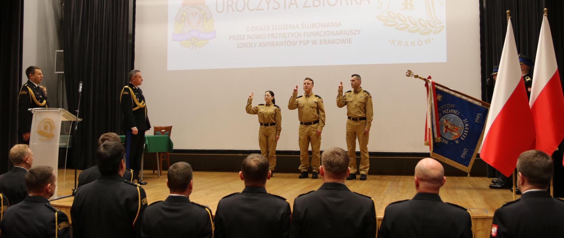 Na scenie Auli SA PSP w Krakowie, w ubraniu służbowym, z uniesionymi w geście ślubowania stoi troje nowo przyjętych funkcjonariuszy. po lewej stronie stoi Komendant Szkoły nadbryg. Piotr Filipek. Z prawej strony poczet sztandarowy SAPSP w Krakowie