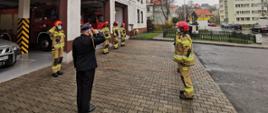 Zdjęcie przedstawia strażaków podczas uroczystej zmiany służby na placu zewnętrznym Jednostki Ratowniczo-Gaśniczej. Strażacy stojący w szeregu naprzeciw siebie. Zmiany służbowej dokonuje dowódca Jednostki Ratowniczo-Gaśniczej. 