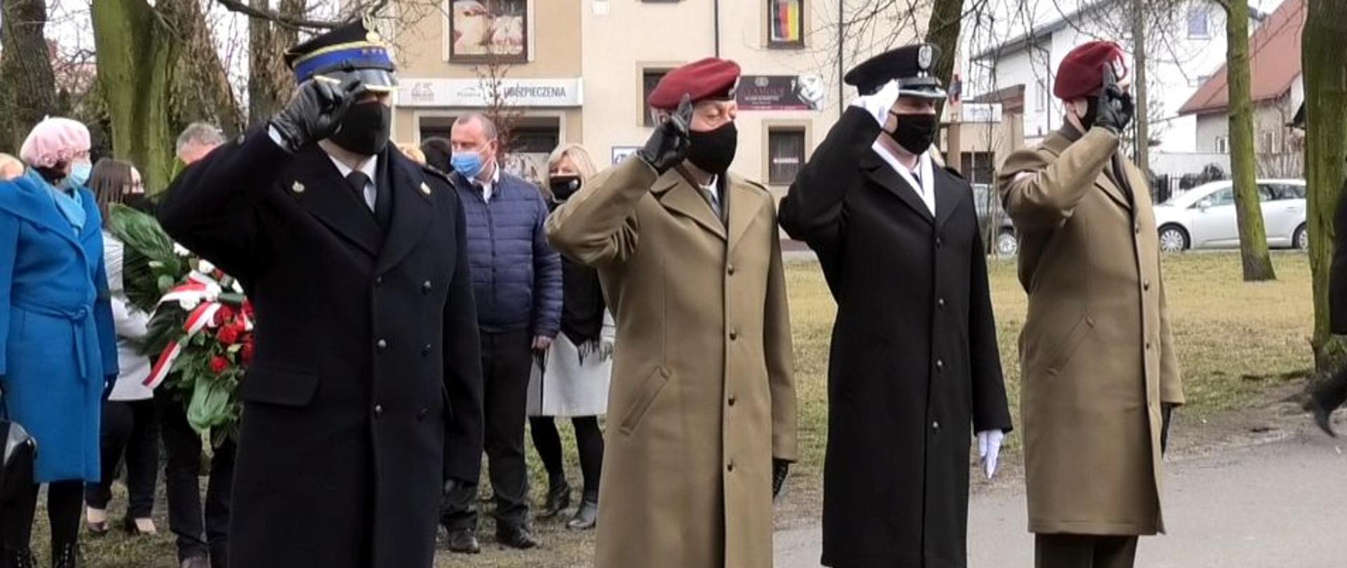 Strażak i trzech żołnierzy w mundurach wyjściowych stoją jeden obok drugiego. Wszyscy salutują. Za nimi stoją ludzie w ubraniach zimowych. Trzymają w rekach kwiaty. W tle widać budynki, drzewa i samochody.
