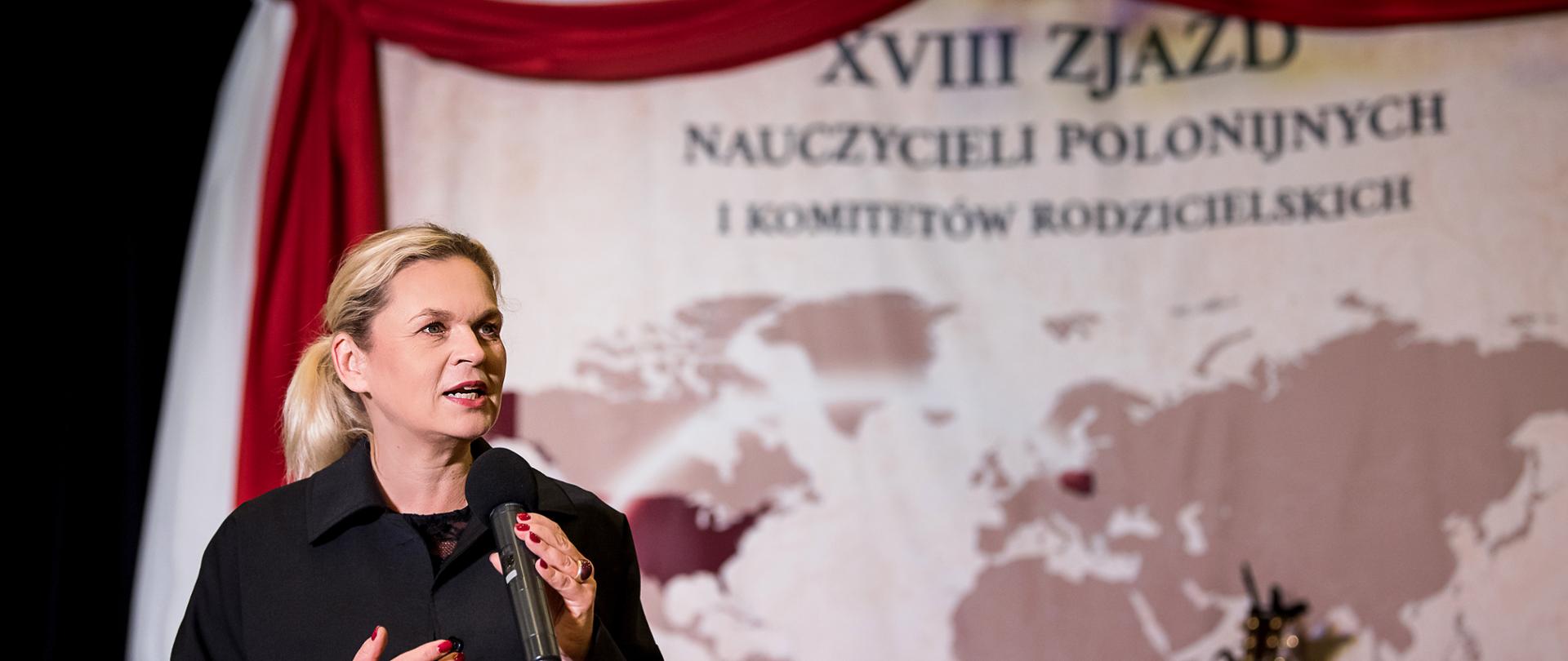 Minister Barbara Nowacka przemawia z mikrofonem na scenie na tle mapy świata i napisu XVIII Zjazd Nauczycieli Polonijnych i Komitetów Rodzicielskich 