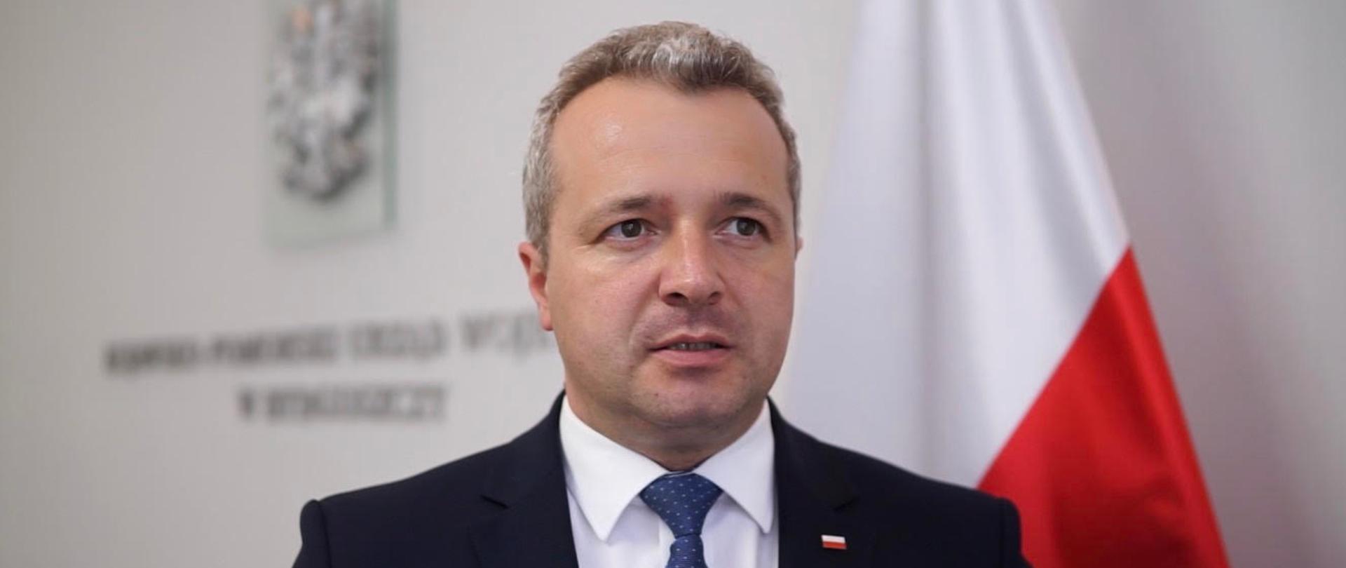 Wojewoda kujawsko-pomorski Mikołaj Bogdanowicz podczas wypowiedzi o aktualnej sytuacji epidemicznej w regionie, w tle biało-czerwona flaga