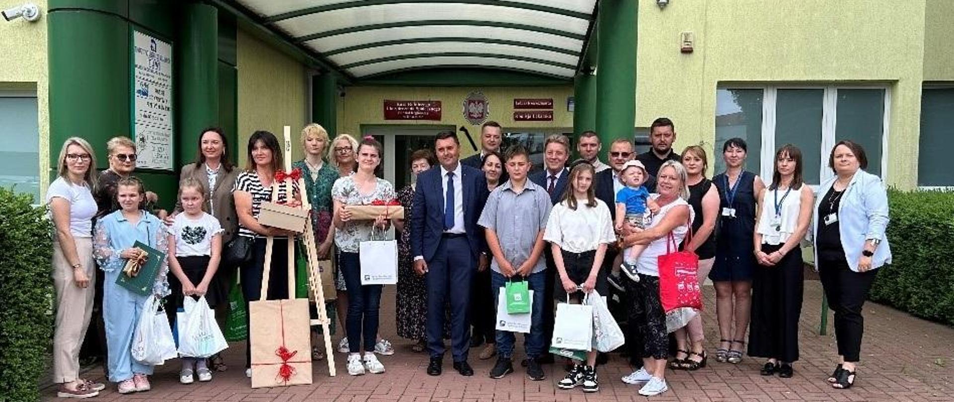 Grupa kilkudziesięciu osób stoi przed budynkiem. Dzieci trzymają w rękach torby i pudełka z nagrodami. 