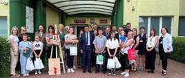 Grupa kilkudziesięciu osób stoi przed budynkiem. Dzieci trzymają w rękach torby i pudełka z nagrodami. 