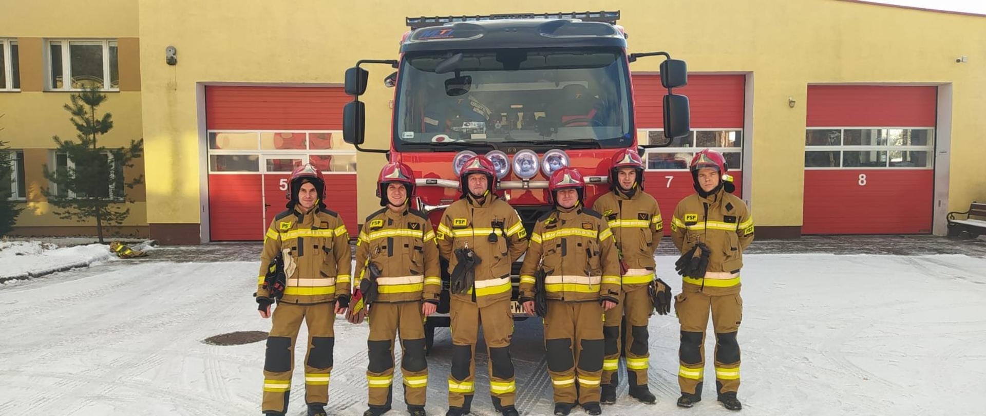 Zmiana służbowa ubrana w piaskowe ubranie specjalne oraz czerwony hełm, na tle nowego samochodu ratowniczo-gaśniczego wykonanego na podwoziu Renault. Samochód koloru czerwonego z niebieskimi światłami ostrzegawczymi oraz srebrnymi roletami. Samochód ustawiony na placu komendy, w tle budynki Komendy Powiatowej PSP w Kętrzynie.