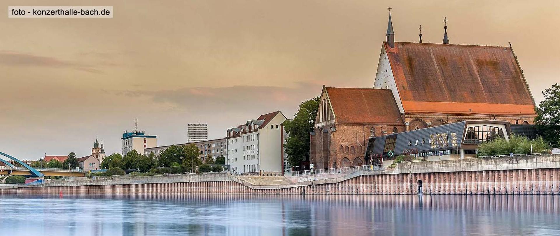 Zdjęcie przedstawia budynek filharmonii wraz z otaczającymi go innymi budynkami. Zdjęcie wykonane ze strony Słubic stąd na pierwszym planie w dole zdjęcia widoczna rzeka Odra.
