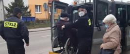 Zdjęcie przedstawia przychodnie zdrowia (punktem szczepień przeciwko COVID-19). Strażak pomaga zejść starszemu mężczyźnie na wózku W tle zdjęcia widzimy drzewa oraz samochody. 